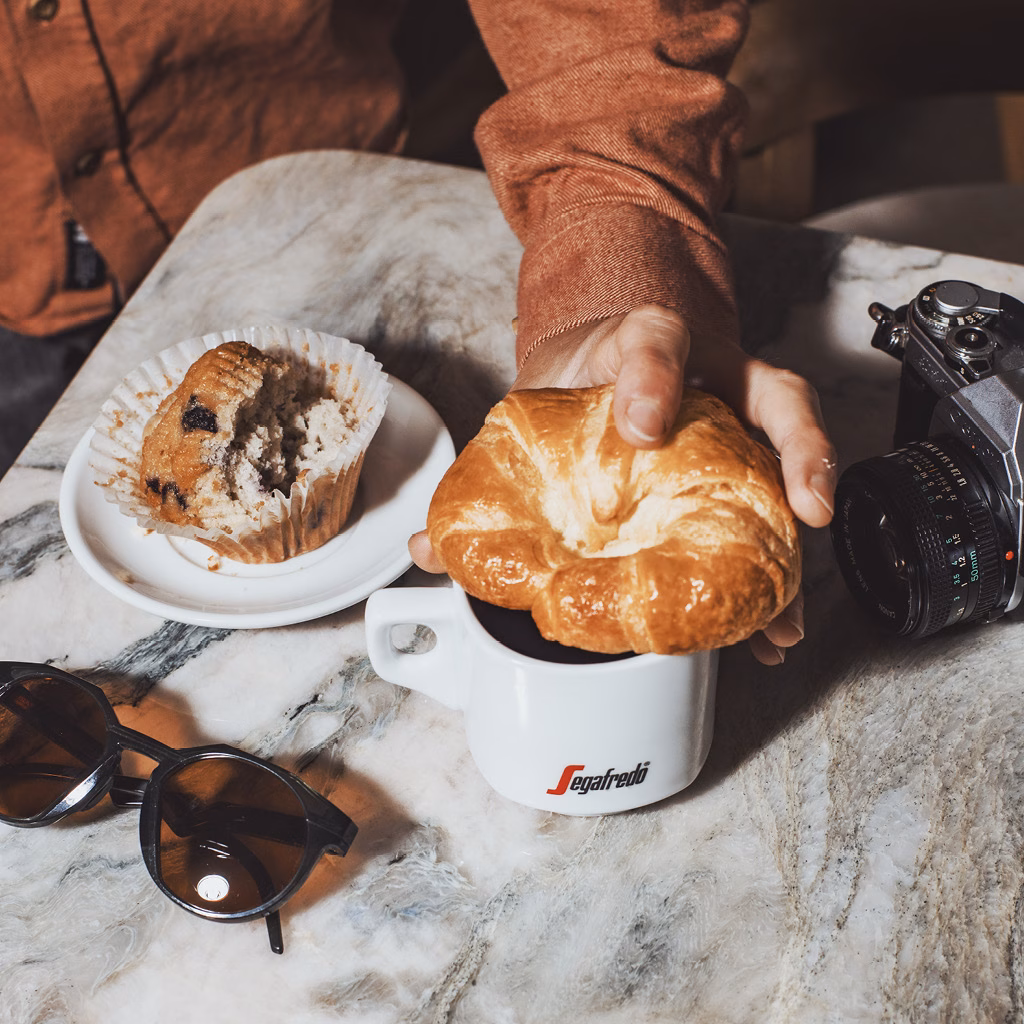 Hand, die Croissant h&auml;lt, Kaffeetasse und Teller mit Muffin